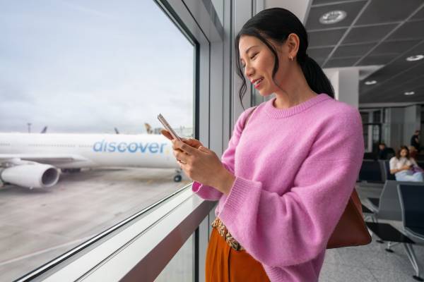 Eine Frau am Gate schaut auf ihr Handy. Im Hintergrund steht ein Discover Airlines Flugzeug.